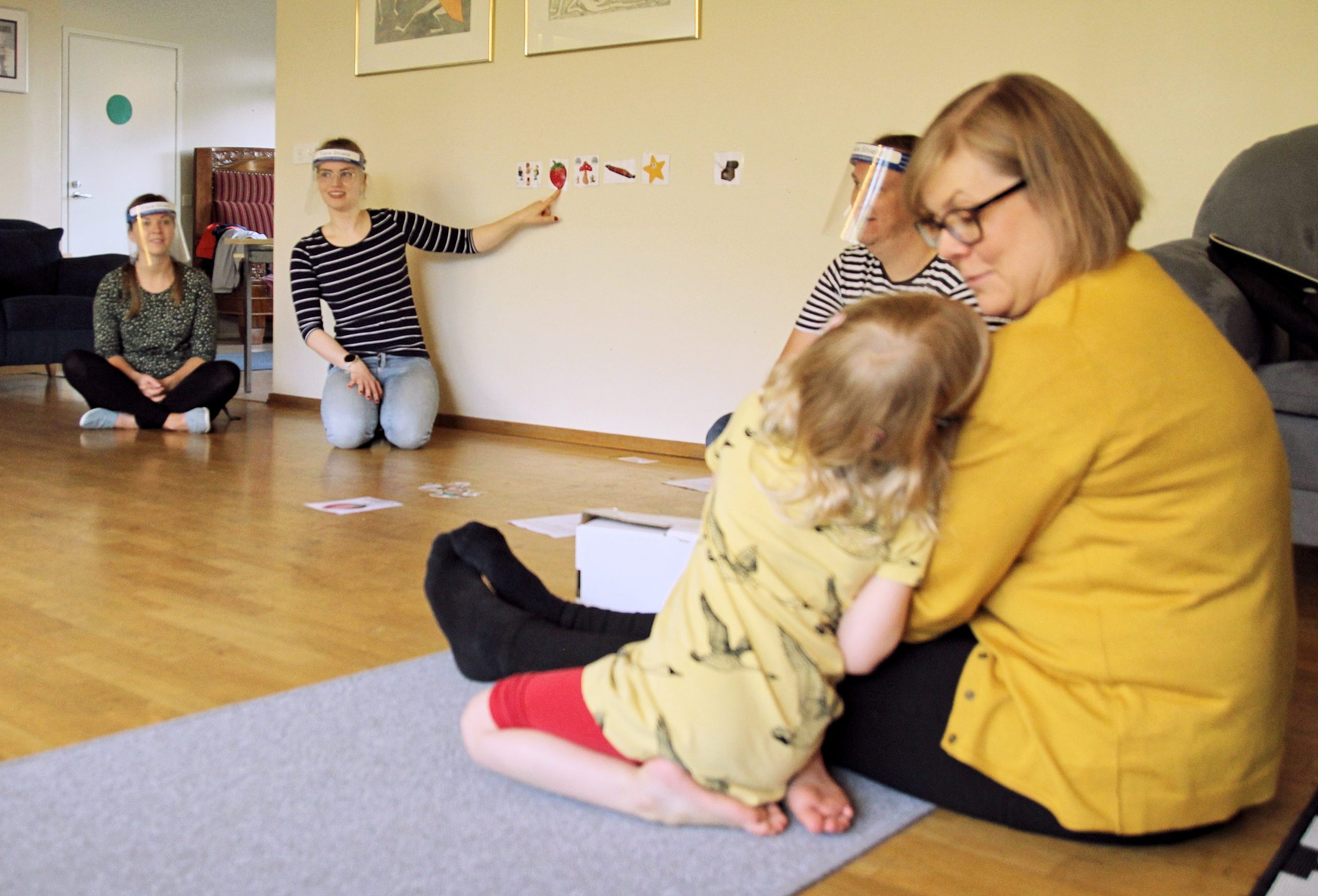 Kuvassa äiti lapsensa kanssa istumassa etualalla, taustalla näkyy visiiriä käyttävä CIsumusan ohjaaja osoittamassa seinällä olevaa kuvaa.